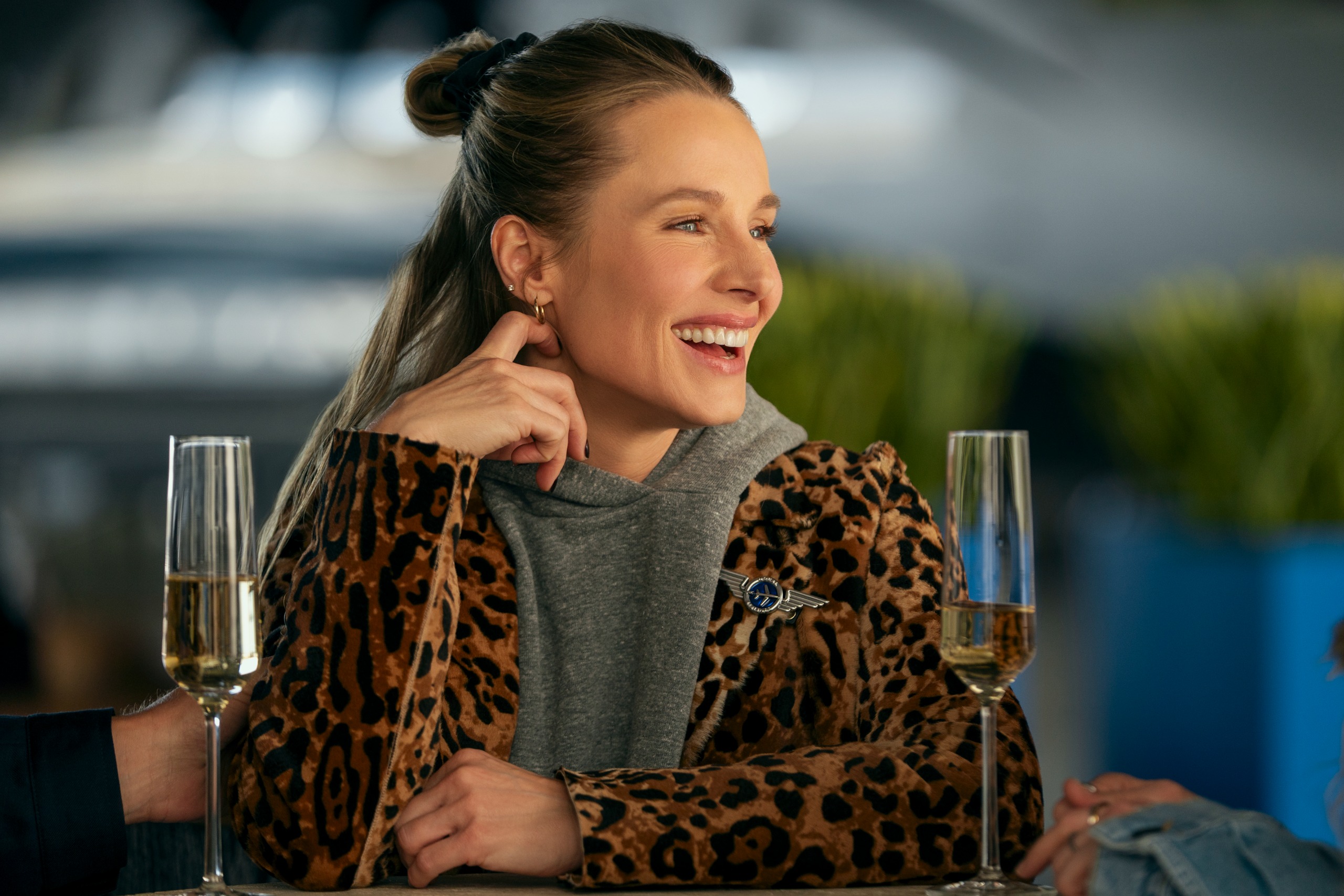 A woman (Kristen Bell as Joanne) wears a leopard-print coat while sitting at an outdoor table in front of wine flutes, in #039;Nobody Wants This#039; season 3.