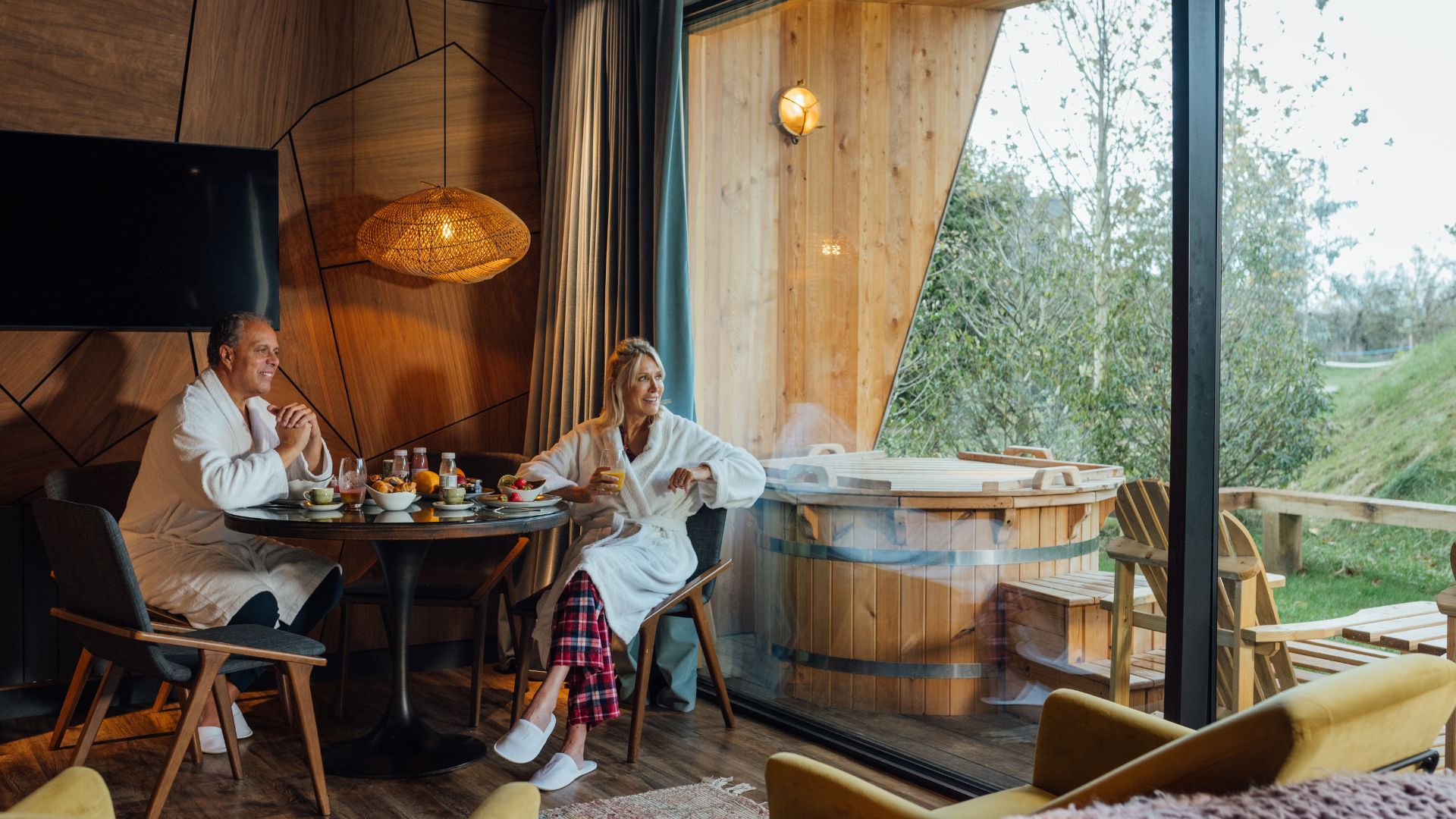 A medium close up side view of a mature couple who have eaten a healthy breakfast whilst enjoying a luxury log cabin get away in Newton By The Sea in the North East of England. They are having a lazy morning and lounging in bathrobes. They are looking forward to using the hot tub on the balcony to relax.