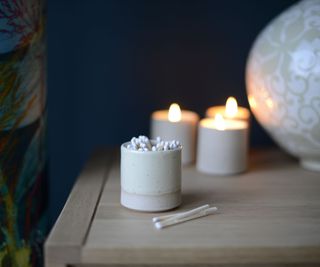 A cream ceramic pot holding white tipped matches. In front of three burning candles in matching containers.