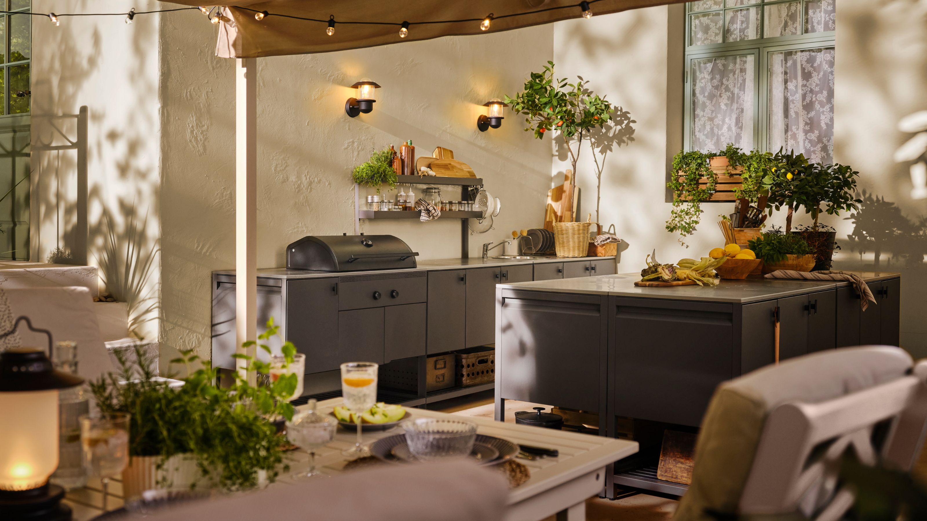 Image of a large, gray outdoor kitchen. There is a row of cabinets and countertops with a grill and a large matching island. In the foreground, there is a table and chairs with plates and cups on it.