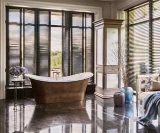 copper coated freestanding bath in large room with floor to ceiling shutters, tall wooden cupboard and tiled floor