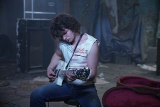 Steve Jones (Toby Wallace) sits in a dirty, decrepit room strumming a guitar, with the open case next to him. He's wearing jeans and a white sleeveless top designed by Vivienne Westwood, reading "you're gonna wake up one morning and know what side of the bed you've been lying on"