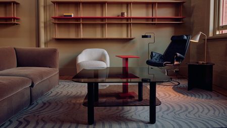 Image of a modern living room in a taupe color with a wavy patterned gray rug. There is a black glass coffee table, a dark taupe sofa, and red shelving on the walls.