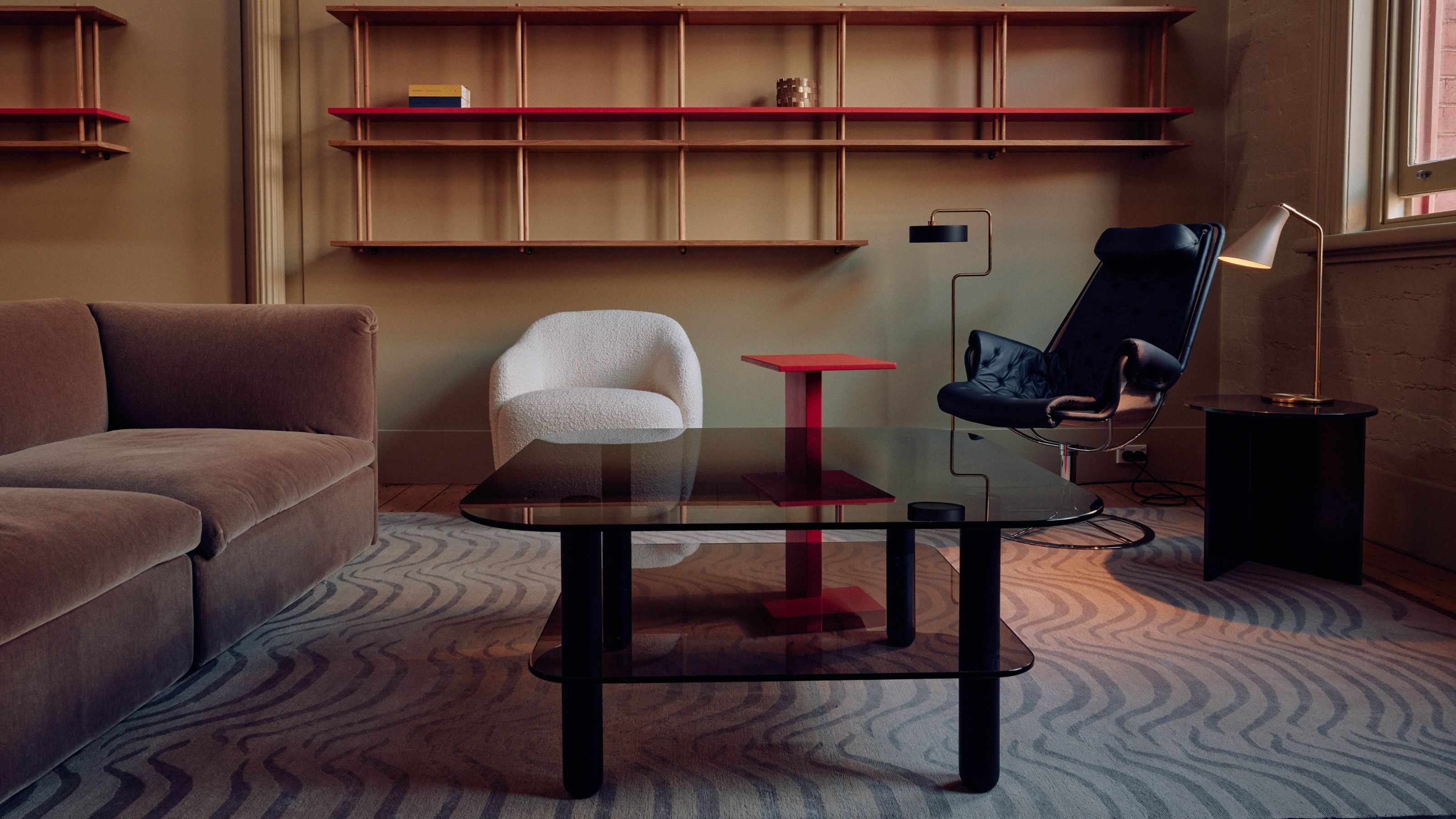 Image of a modern living room in a taupe color with a wavy patterned gray rug. There is a black glass coffee table, a dark taupe sofa, and red shelving on the walls.