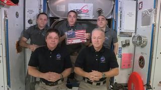 NASA astronaut Chris Cassidy (center) displays an American flag that Demo-2 astronauts Bob Behnken and Doug Hurley (bottom row from left) will return to Earth aboard their SpaceX Crew Dragon spacecraft. The flag was a target for the first commercial crew mission to reach the station.