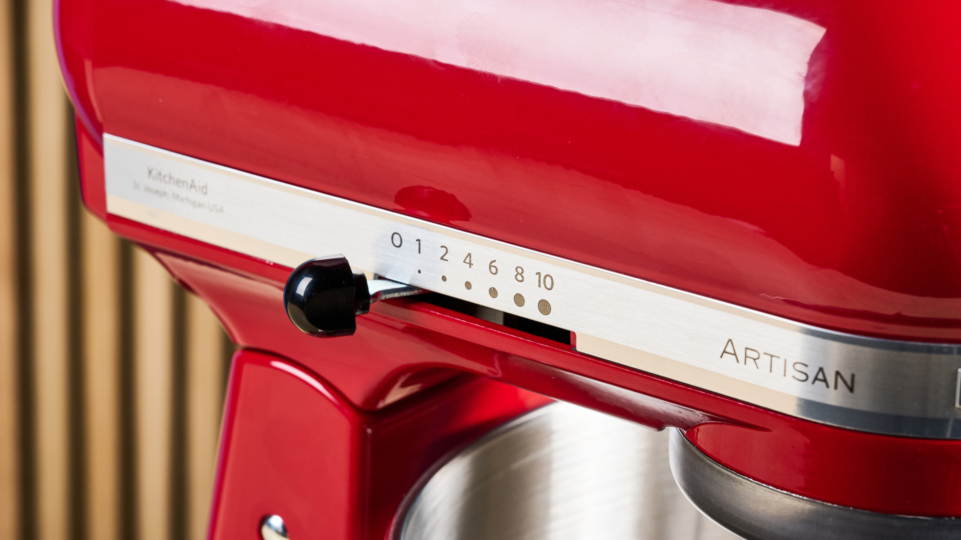 photograph of the kitchenaid artisan stand mixer tilt head in cherry red, with the mixing bowl, paddle, dough hook, and whisk attachments, photographed against a blue background