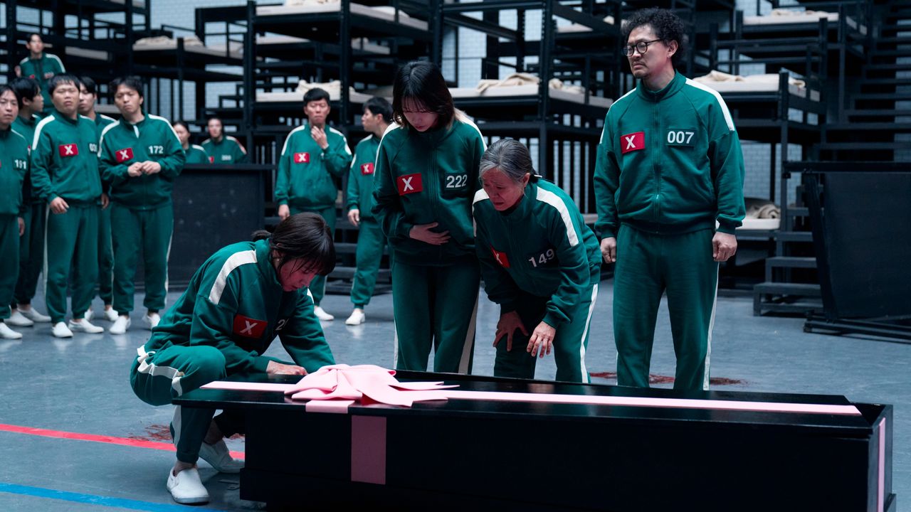 Squid Game season 3 players in a big room all wearing matching green and white tracksuits standing over a black coffin with a pink bow and looking inside at something we can't see