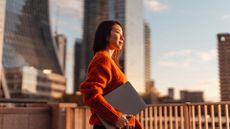 A businesswoman in an orange jumper holding a laptop, looking to the right of frame. In the background, a cityscape at sunset can be seen, with orange sunlight making the businesswoman's face glow.
