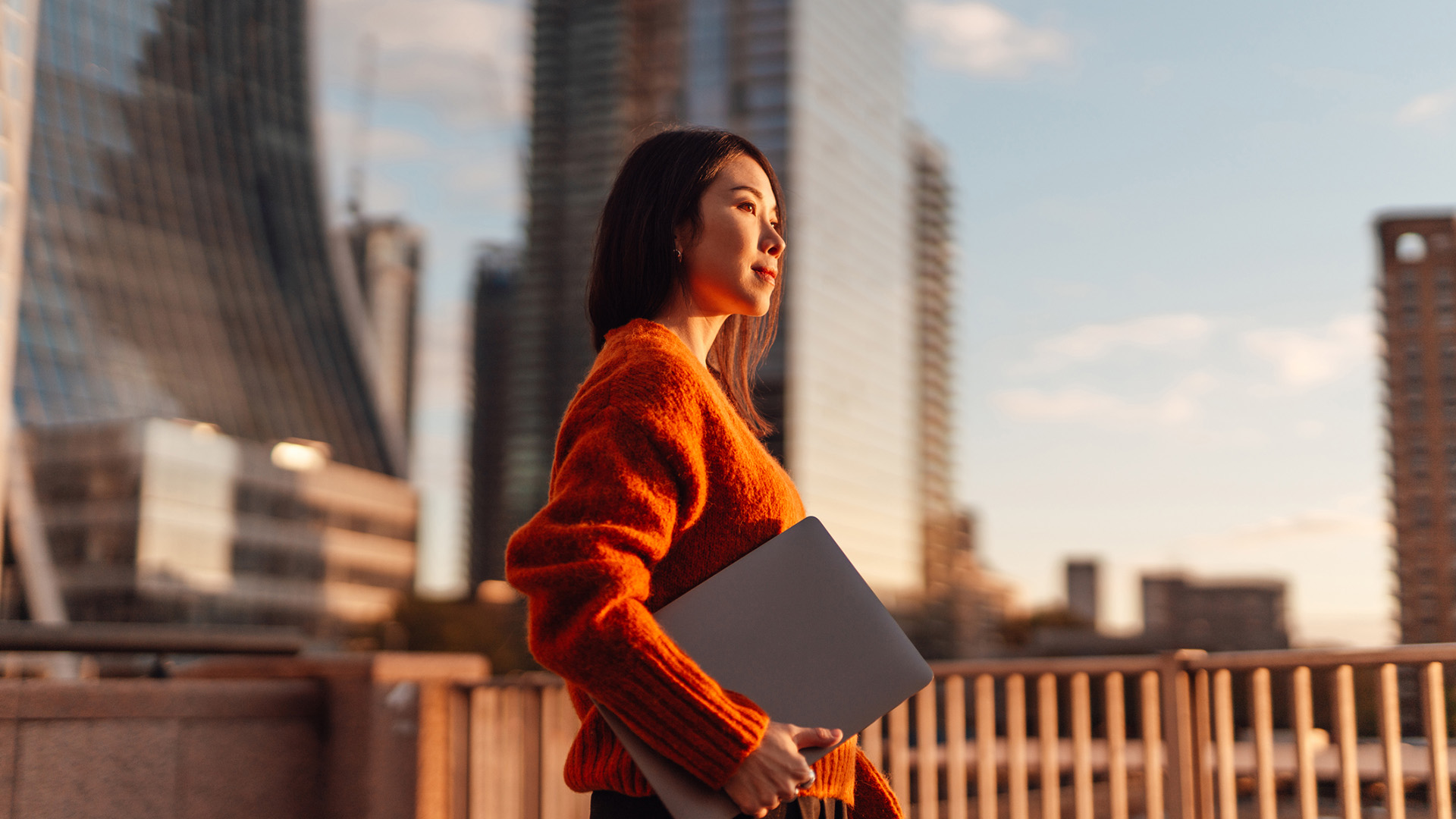 A businesswoman in an orange jumper holding a laptop, looking to the right of frame. In the background, a cityscape at sunset can be seen, with orange sunlight making the businesswoman's face glow.