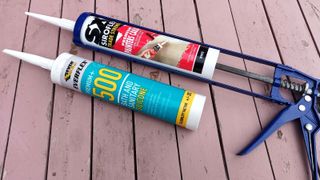 Two caulk cartridges side by side with on ein caulking gun on pink wooden table