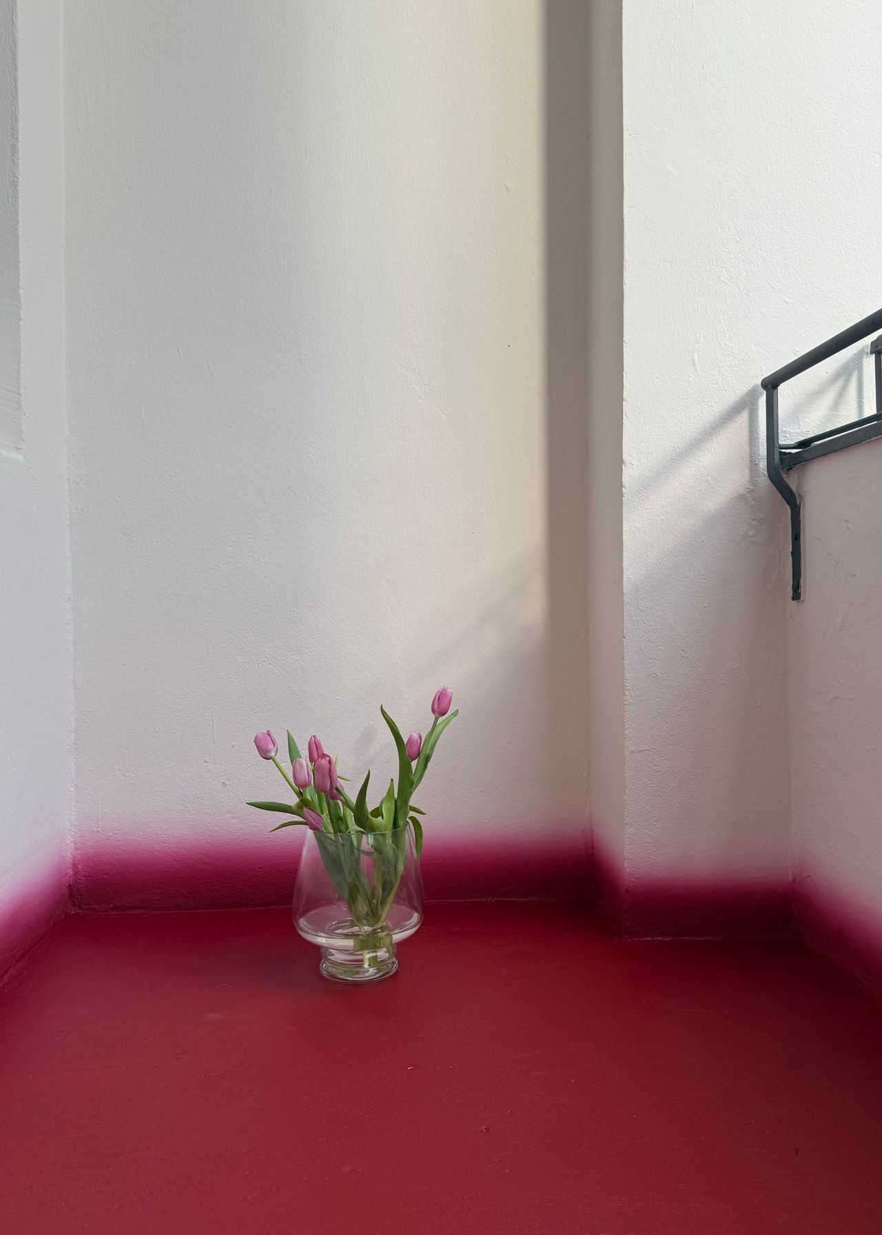 Image of a clear vase of pink tulips on a white balcony with a red floor that has a gradient skirting board.