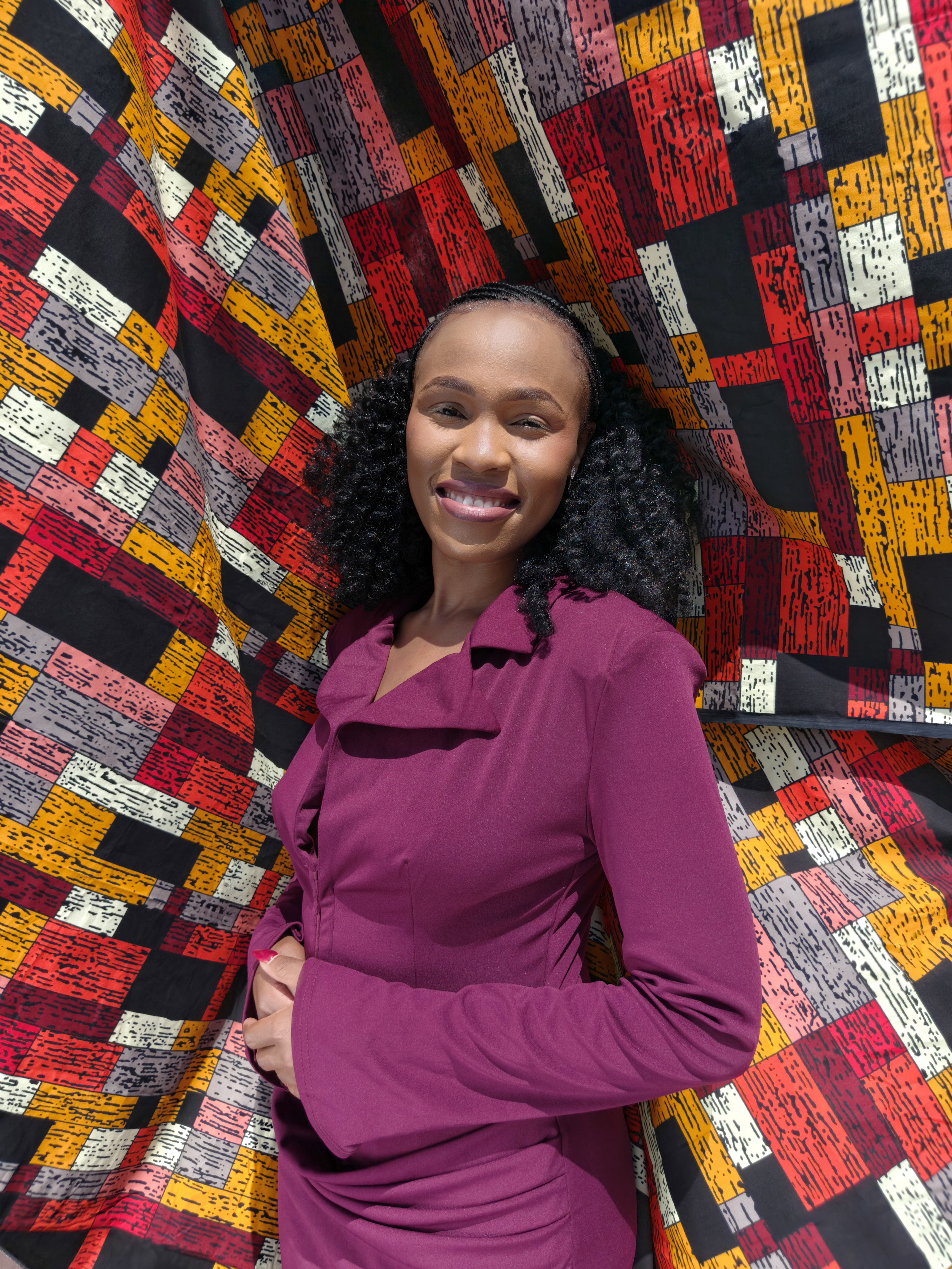 A smiling woman with shoulder-length curly hair, wearing a plum long-sleeved dress, poses in front of draped fabric printed with red, orange, grey and black brick-like blocks.