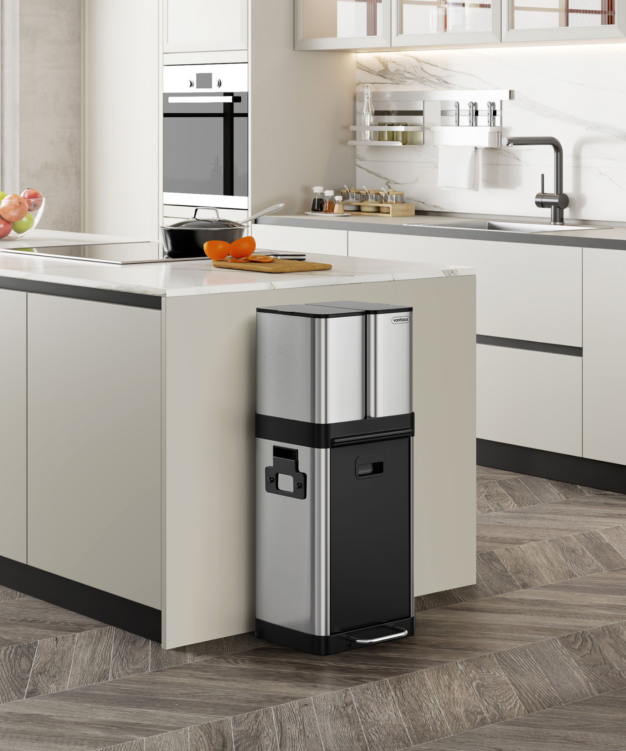 A slim, tall stainless steel and black kitchen bin with dual compartments and a foot pedal, blending into a modern kitchen beside a marble-topped island.