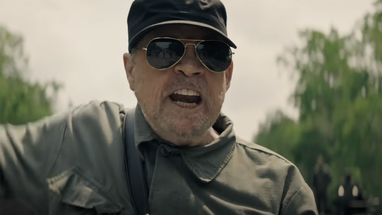Mark Hamill in sunglasses, fatigues and a hat hollering outside in The Long Walk.