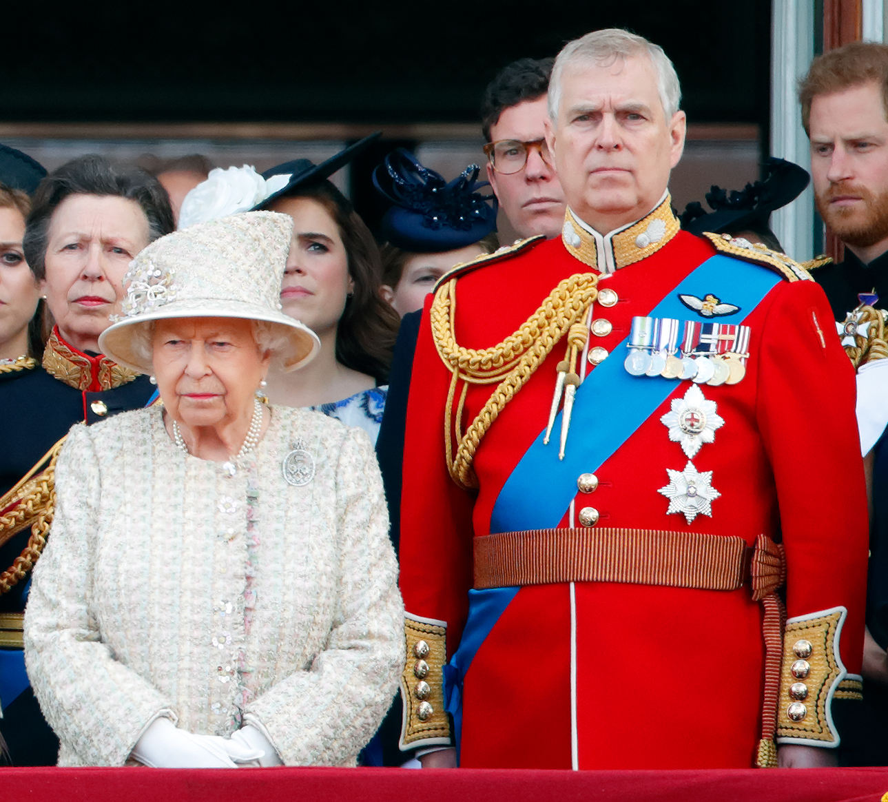 Queen Elizabeth wearing an ivory coat and hat standing next to Andrew Mountbatten-Windsor, in a red military uniform, and other members of the Royal Family on the balcony of Buckingham Palace