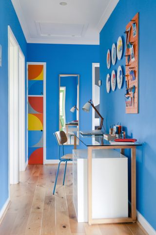 Blue painted hallway with a desk and chair, plates on the wall and, at the end, a cupboard door painted with curved geometric designs in reds, oranges, yellows and blues