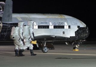Here, the X-37B space plane is seen in profile as post-landing work continues. The logos of Boeing and the Air Force are visible on the reusable spacecraft's hull. They appear between lines that outline the X-37B's payload bay, which is about the size of