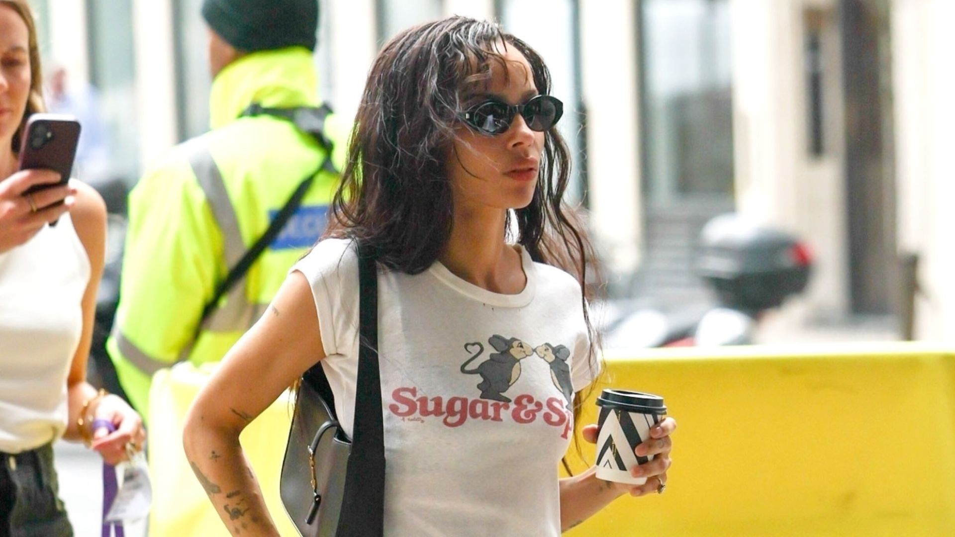 Zoe Kravitz walks outside holding a coffee wearing a white graphic t-shirt, black trousers and black heeled peep-toe shoes.