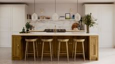 A kitchen with a wooden kitchen island and stools with a marble countertop, three glass pendant lights, and a marble splashback with shelves on top of it