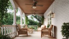 Photograph of porch of white-brick house, with four rattan chairs, two ceiling light fans and flowers. The porch is surrounded by greenery and trees.