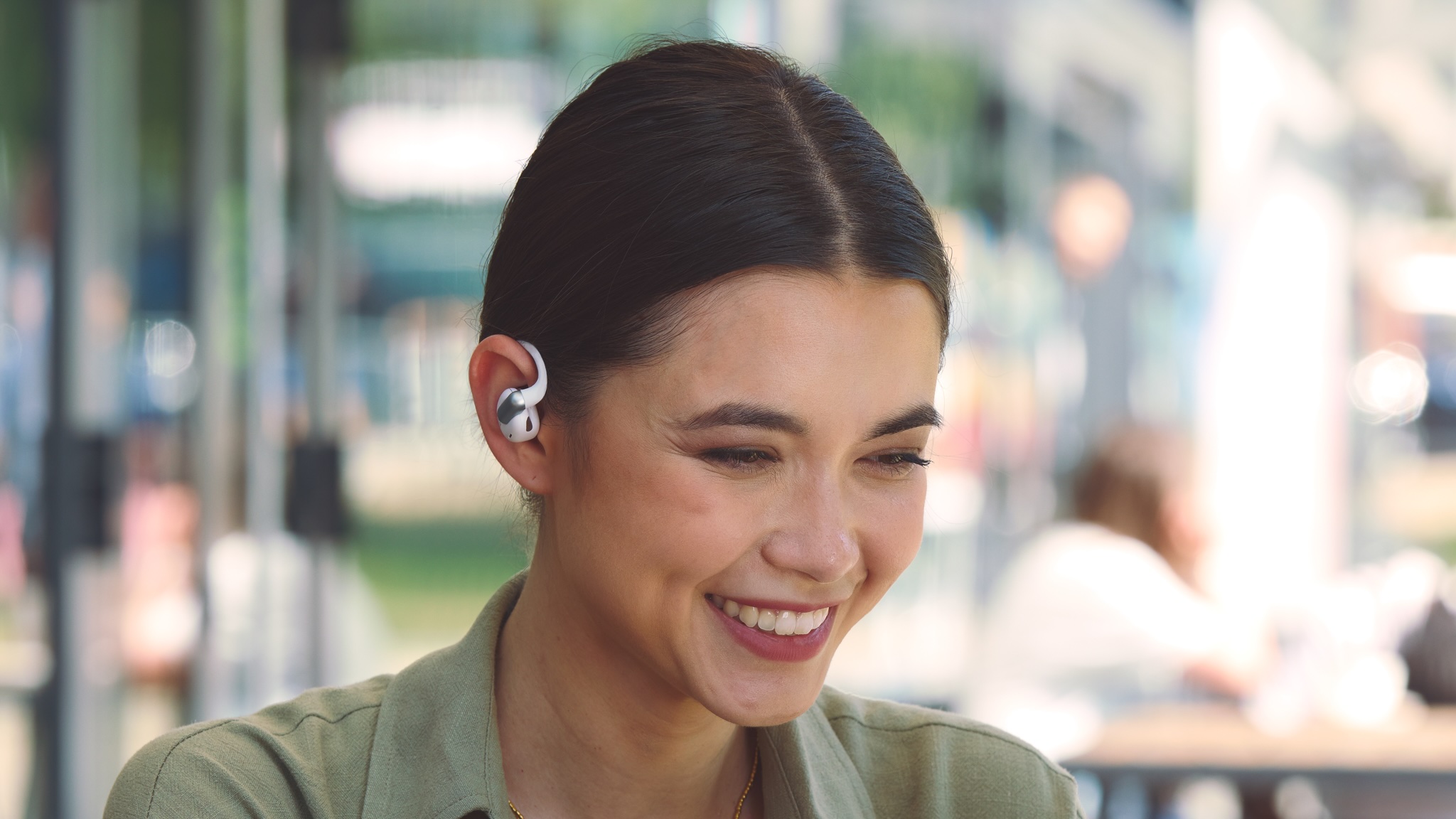 A smiling young woman with a sage green top wearing JBL Sense Pro over-ear headphones in white in what looks like a café - the background is blurry