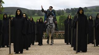 Host Alan Cumming welcomes his new celeb guests on horseback, donning his traditional Scottish attire and surrounded by creepy figures in black cloaks and wearing gold masks, in The Traitors US Season 3. 