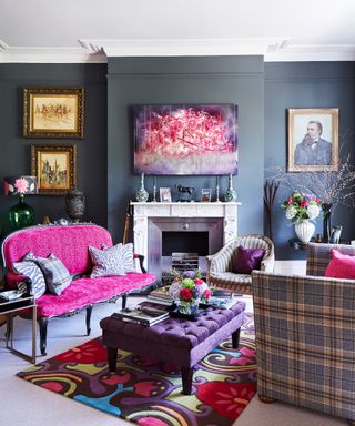 A blue-grey painted living room with hot pink sofa, colorful pink, purple and green artwork and rug, and a purple footstool, with gilt framed paintings on the wall.