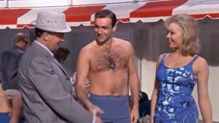 Sean Connery as James Bond in a bathing suit greeting a man in a suit while standing next to a woman in a blue bathing suit.