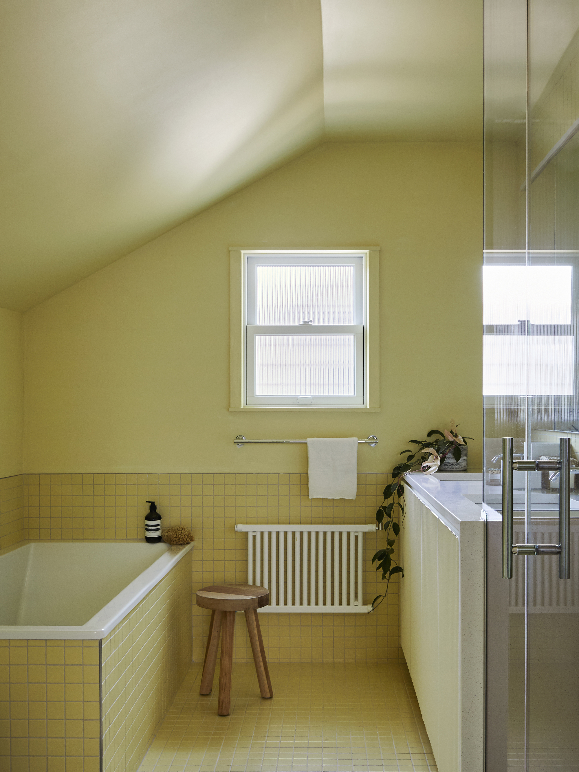 yellow bathroom with wall paint and yellow mosaic tiles, a built-in yellow bath with a timber stool next to it, radiator, towel rail, small winder, and white vanity with a potted trailing plant on it