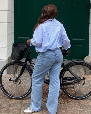 Influencer wears baggy jeans with blue button-down shirt, standing with back to camera in front of black bike.