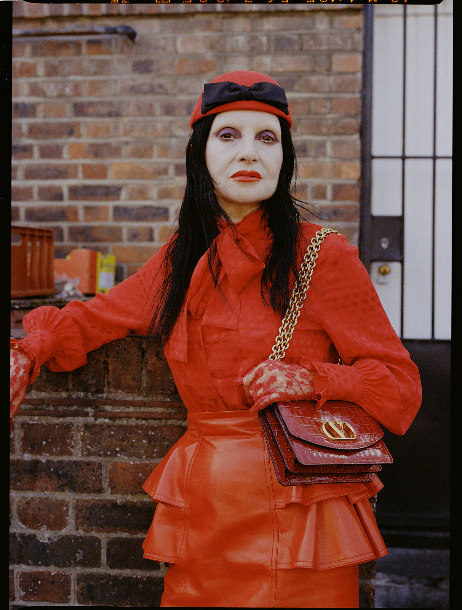 A person in vibrant red attire with black hair leans against a brick wall. They wear a matching red hat and hold a red shoulder bag with a gold chain