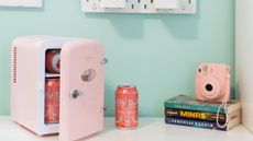 Koolatron 6 Can AC/DC Retro Mini Cooler Personal Mini Fridge Refrigerator in pink in blue room with soda can beside it and a stack of books and a pink polaroid camera