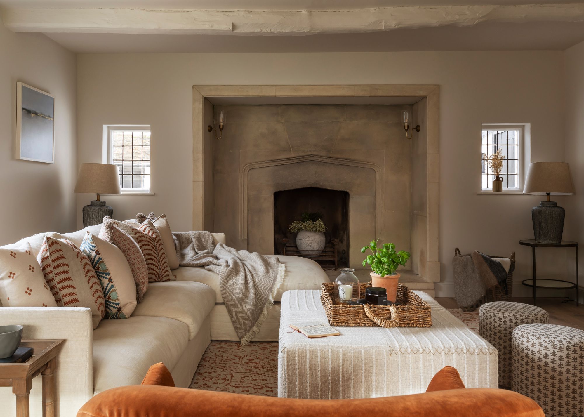 A cottage living room with warm white walls, a ceiling beam, and a large stone fireplace. A cream chaise sofa with layered cushions and a cream striped ottoman.