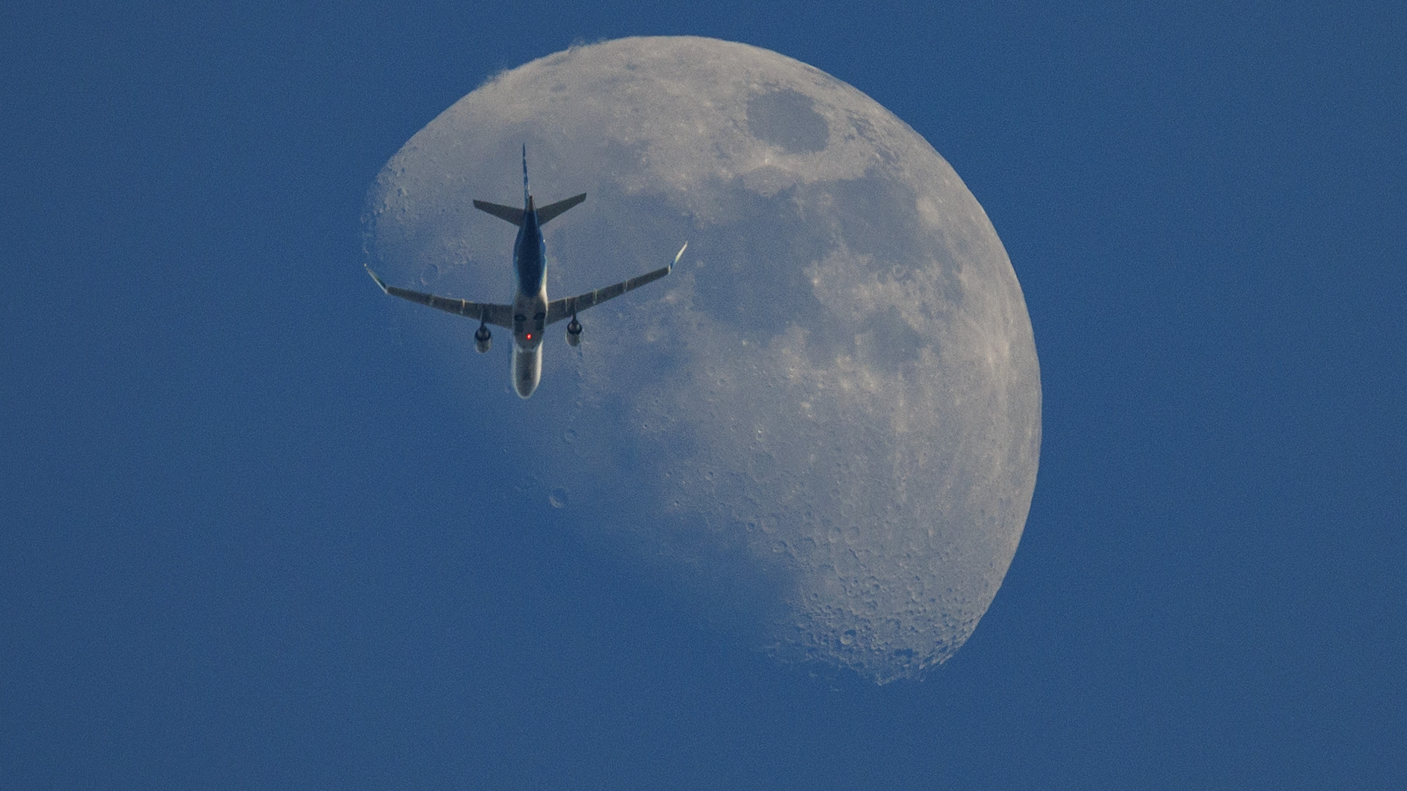 A waxing moon is pictured in the blue daytime sky with its right side lit by sunlight as a commercial jet flies in front of its disk.