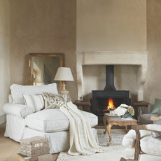 Neutral cosy living room, stone fireplace, lit stove, giant oversize clock face, grey striped armchair, gold gilded coffee table, wood flooring