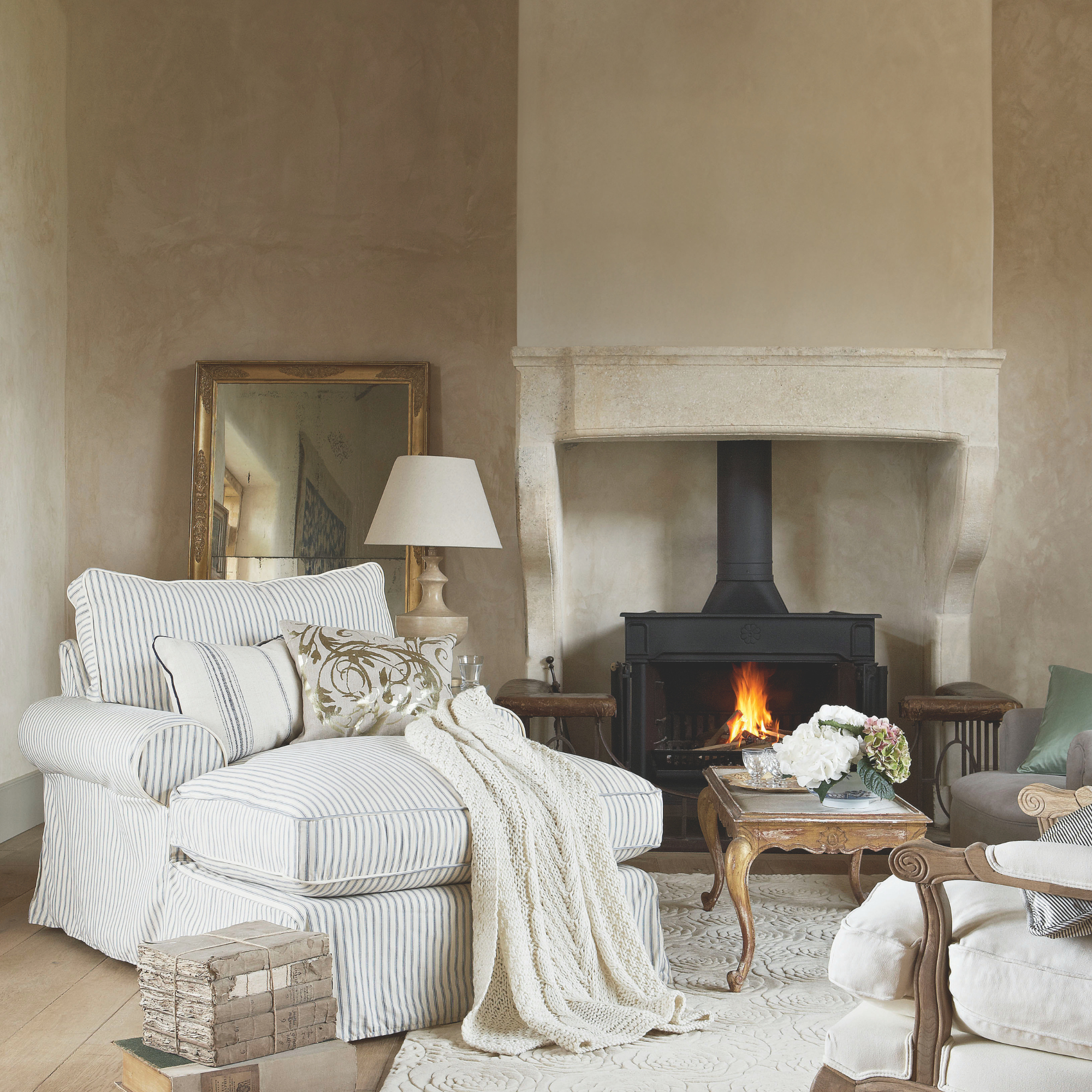 Neutral cosy living room, stone fireplace, lit stove, giant oversize clock face, grey striped armchair, gold gilded coffee table, wood flooring
