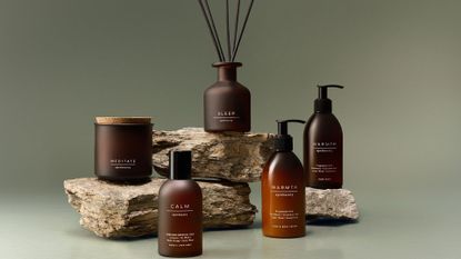 A set of amber vessels in diffuser, candle, room spray, moisturizer, and soap form on stacked rocks against a sage green backdrop