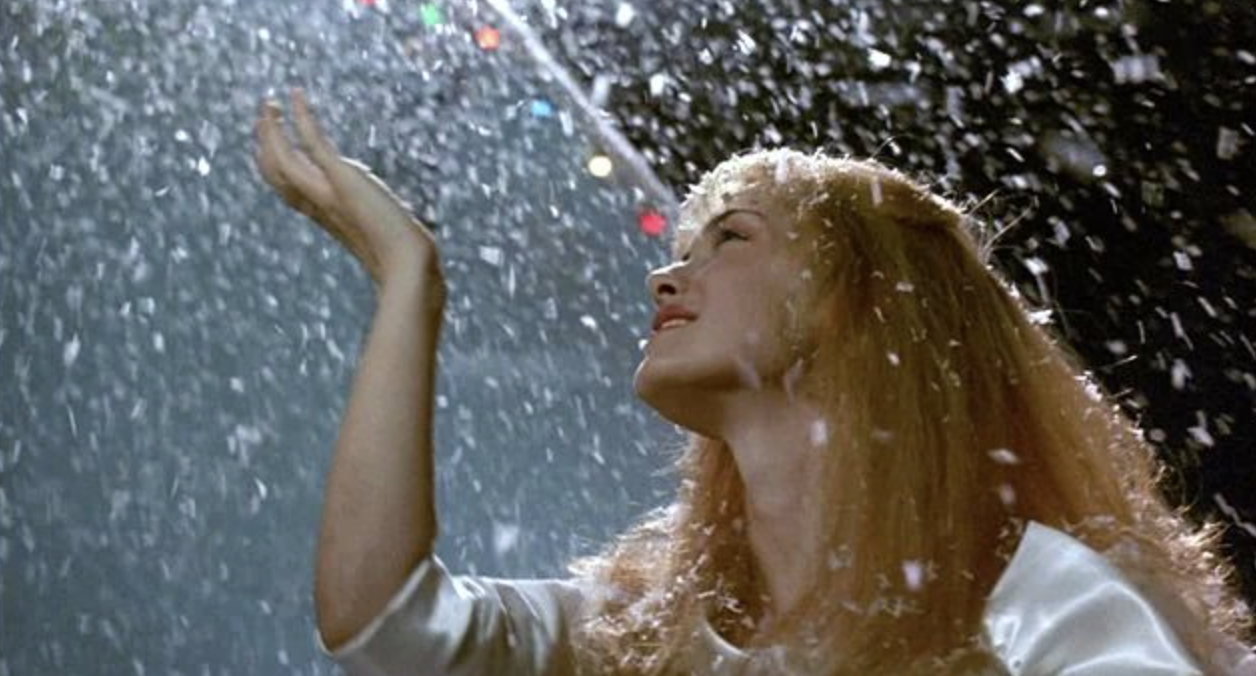 A young woman with long blonde hair looks up with a peaceful expression, reaching her hand out toward falling snow against a dark, festive background.