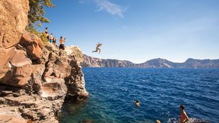 Young people wait on a rock outcrop and swim or jump into the deep blue waters of a mountain lake, Crater Lake, USA