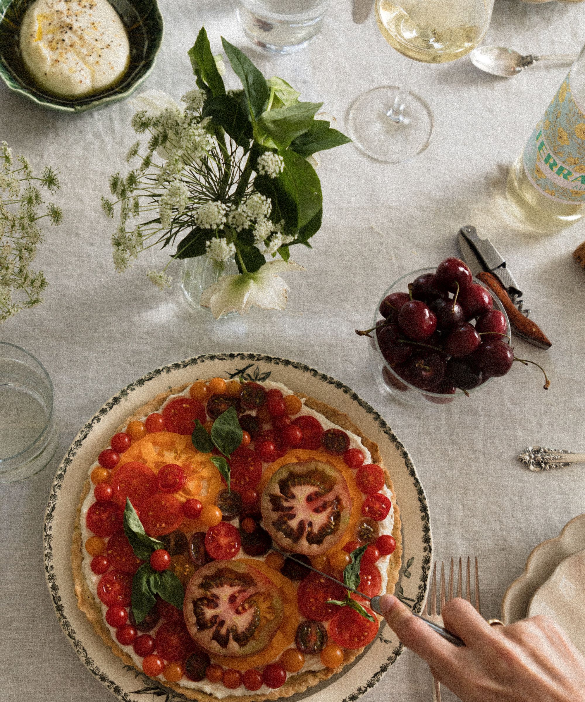 Outdoor tablescape with a linen tablecloth, a bud vase filled with fresh white flowers, a china green and white plate with a tomato tart, a bowl of cherries, and wine glasses