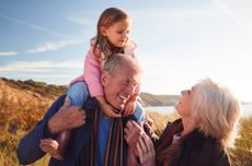 Grandfather Giving Granddaughter Ride On Shoulders As They Walk Through Sand Dunes With Grandmother.
