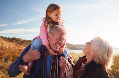 Grandfather Giving Granddaughter Ride On Shoulders As They Walk Through Sand Dunes With Grandmother.