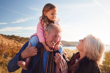 Grandfather Giving Granddaughter Ride On Shoulders As They Walk Through Sand Dunes With Grandmother.
