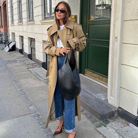 fashion influencer Felicia Akerstrom poses on a sidewalk in Copenhagen wearing black oval sunglasses, a trench coat, white t-shirt, cuffed jeans, black leather shoulder bag, and burgundy slingback heels.