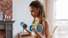 A woman performs a biceps curl with dumbbells in a living room. Her elbows are bent and the light dumbbells are close to her shoulders. In the background we see a red brick wall, a fireplace and candles. 