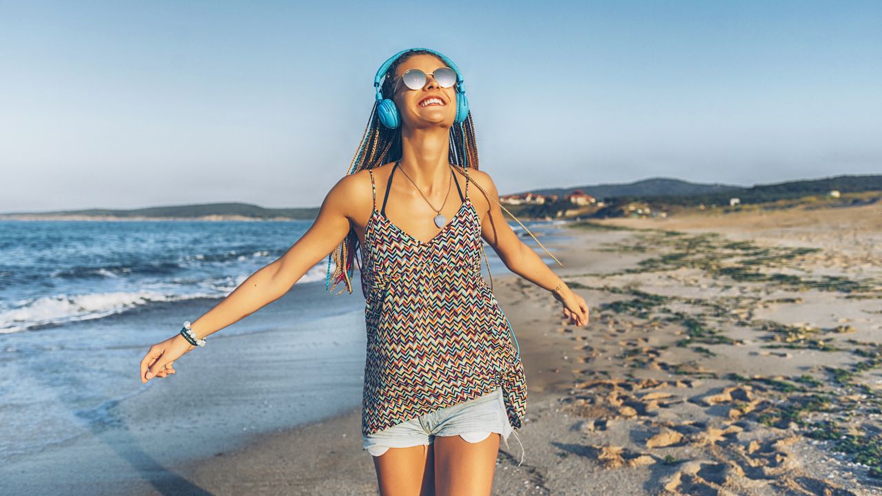 Cheerful young woman with blue headphones listening music on beach.