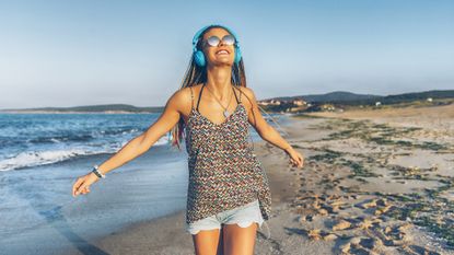 Cheerful young woman with blue headphones listening music on beach.