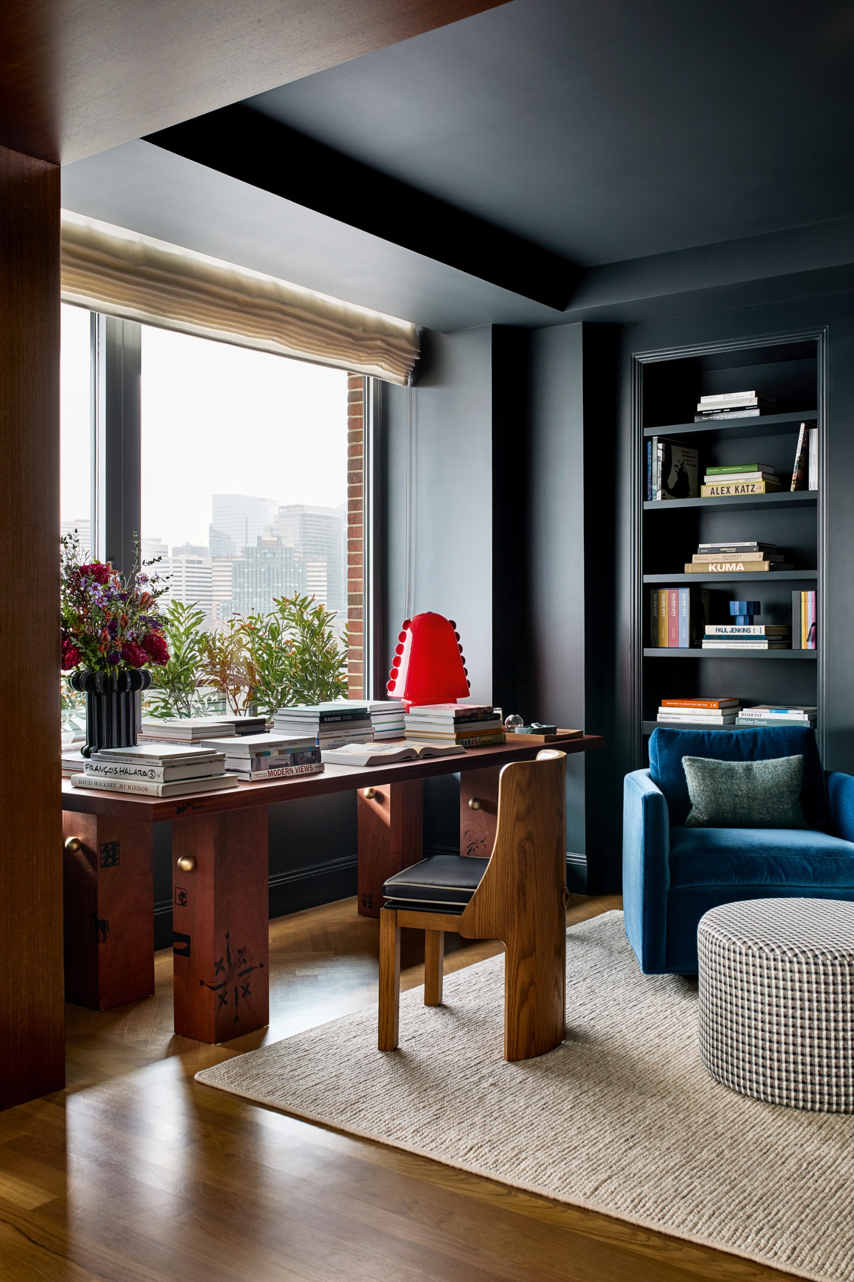Dark blue living room with library table in the corner, topped with books and a red lamp, with a curved wooden chair, blue armchair and beige rug