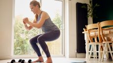 A woman performs a squat at home in her kitchen, wearing leggings and a vest. She is stood on an exercise mat with dumbbells to the side. Behind her is a long wooden table with chairs.
