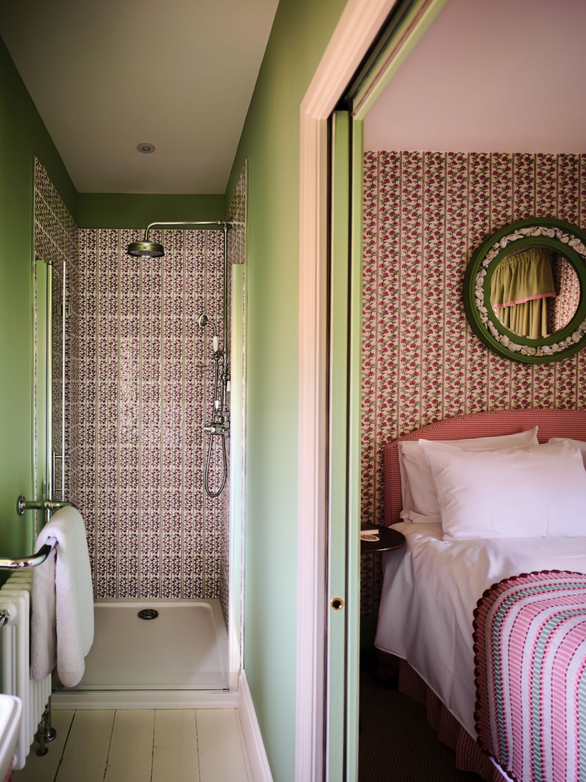 Narrow green en suite with floral striped shower tiles beside a bedroom with matching wallpaper, a pink headboard and a round green mirror.